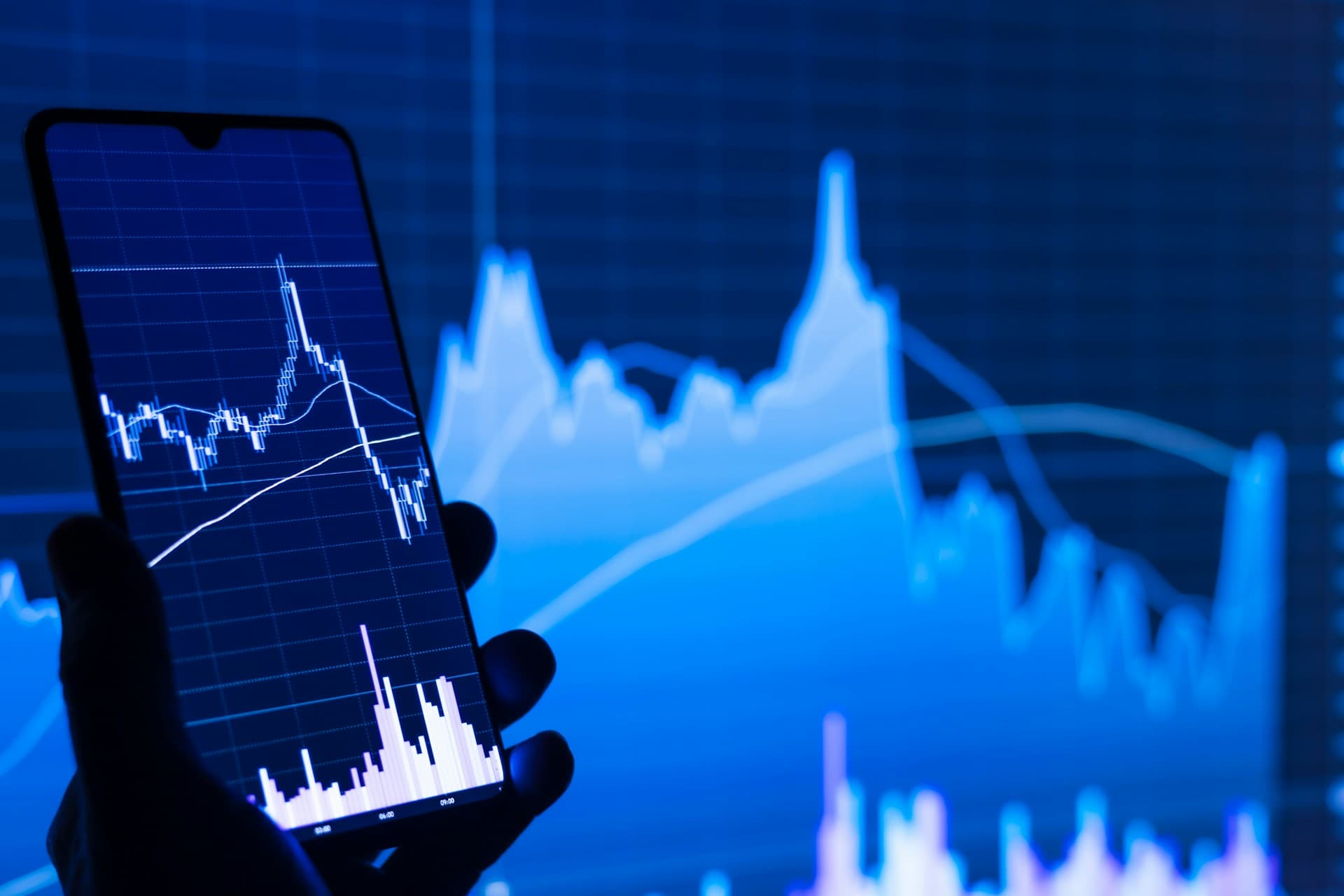 A hand holding a smartphone displaying financial charts, with data graphs in the background.